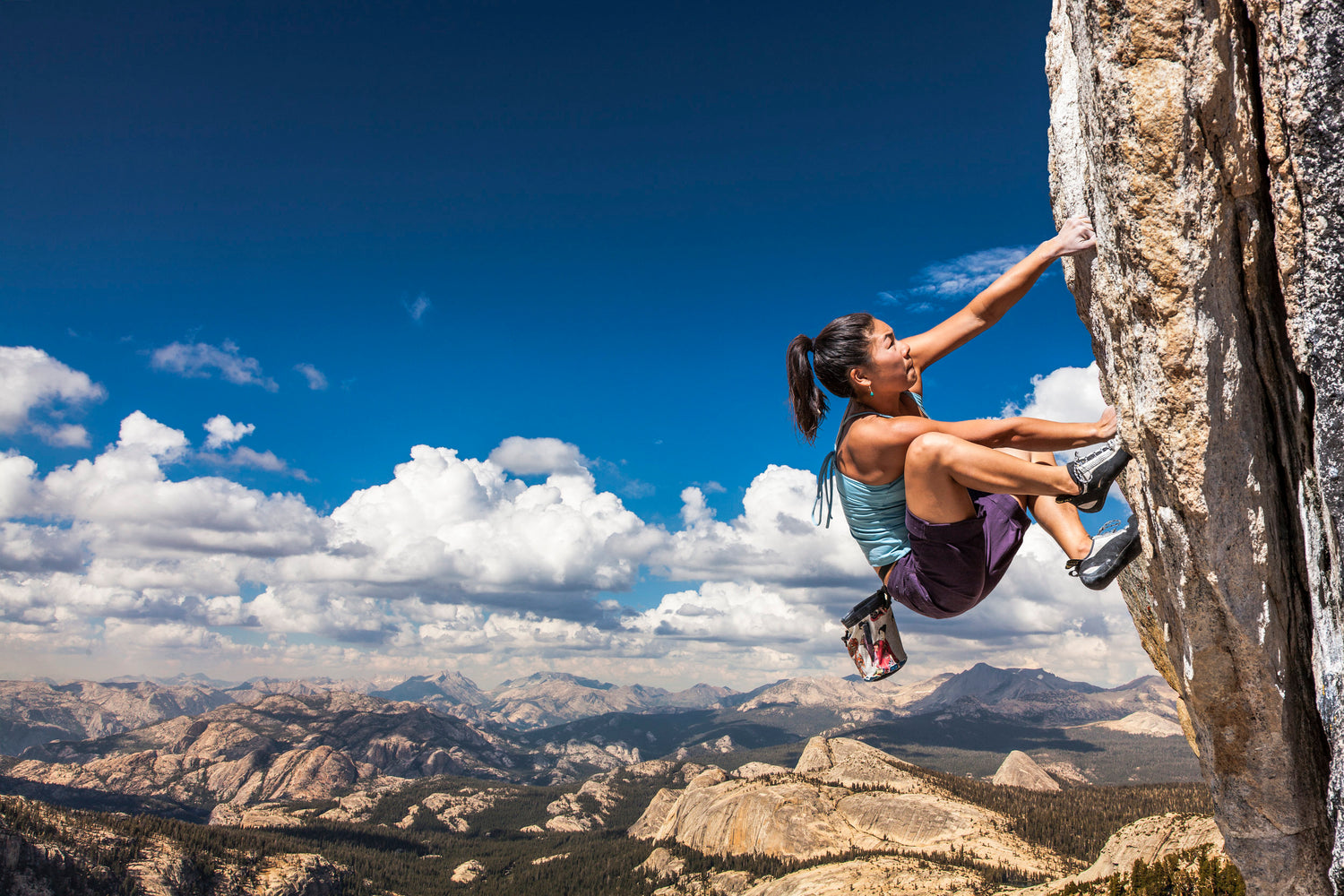 Woman rock climbing in crunched position, ready for powerful move upward, and bouldery landscape in distant background -- elevation implied. Credit: https://www.melaninbasecamp.com/trip-reports/2018/7/31/eight-amazing-asian-pacific-islander-women-climbers